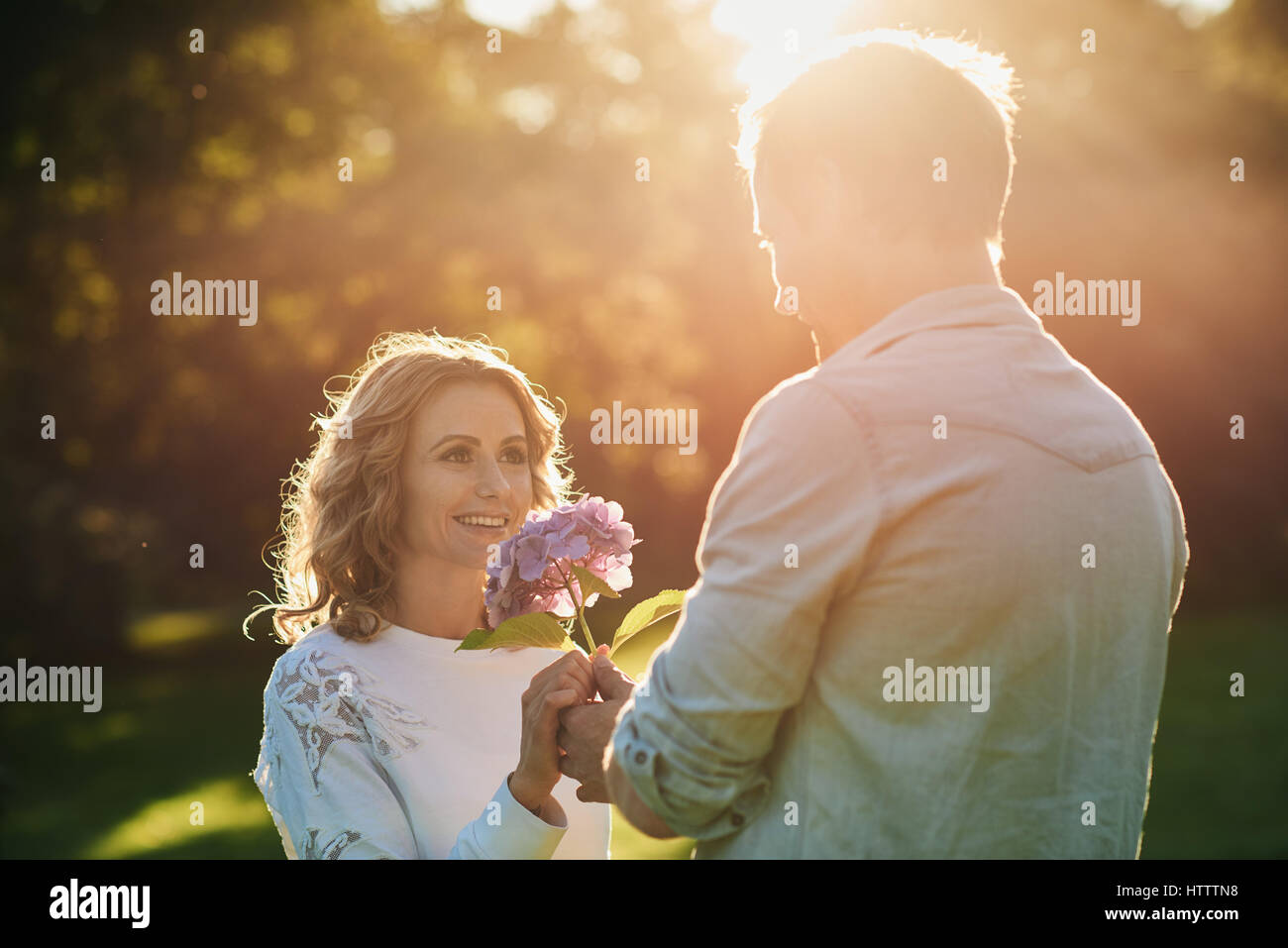 Handsome young man giving his beautiful wife a bouquet of purple wild flowers while enjoying a romantic afternoon in a park in the summer Stock Photo