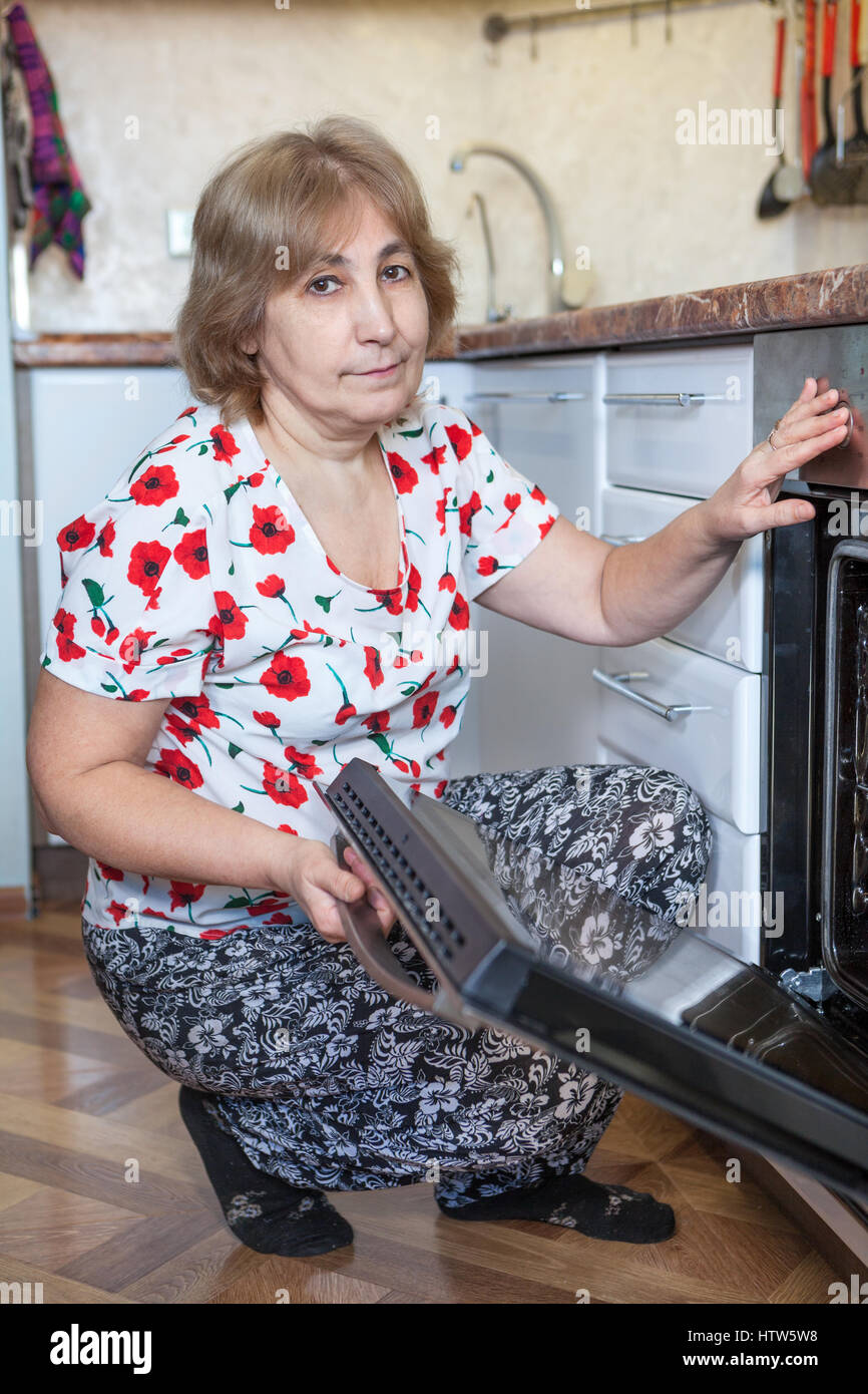 Mature Caucasian woman sitting beside the built-in oven with open door. Domestic kitchen Stock Photo