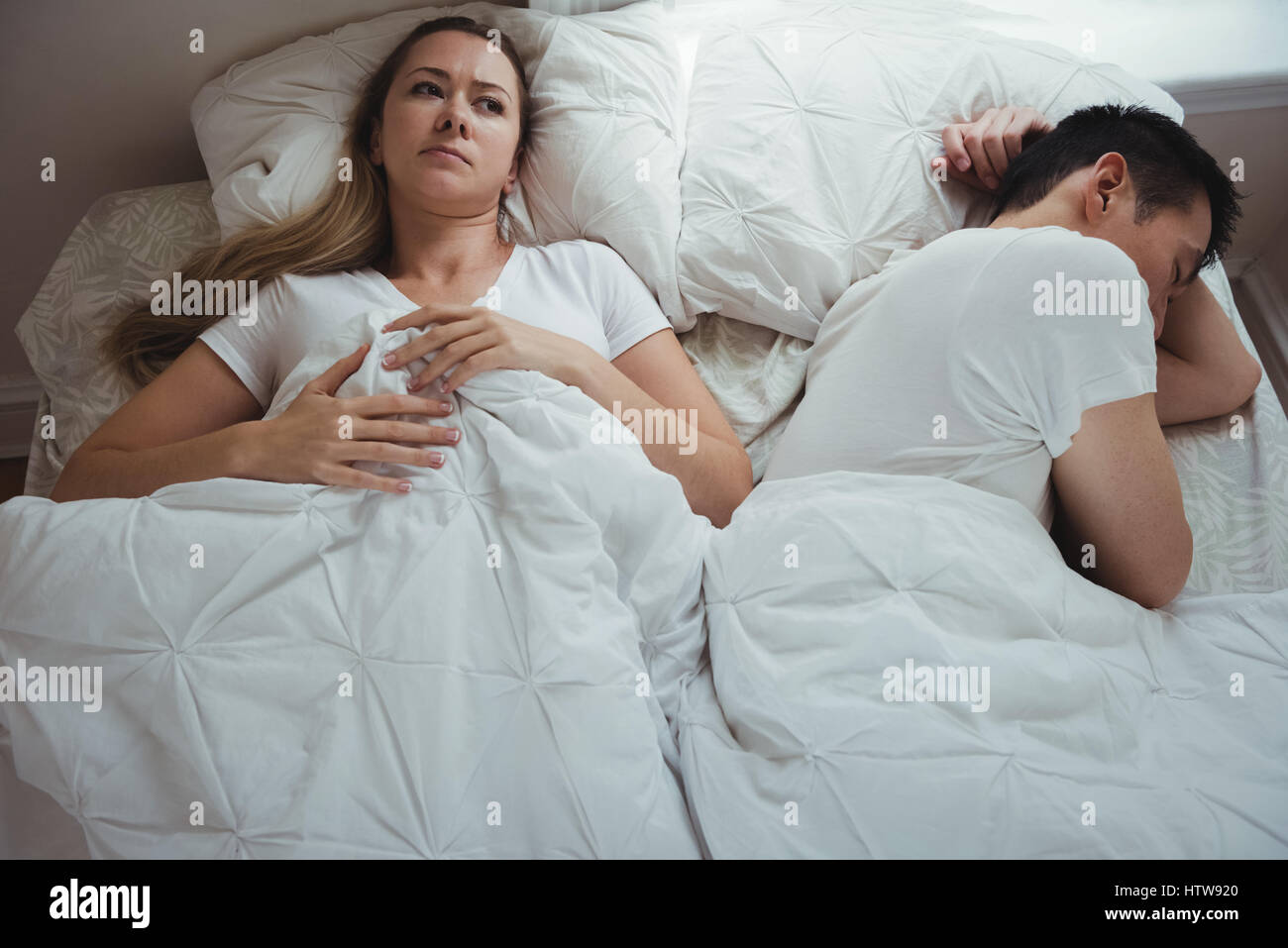 Worried woman lying while man sleeping beside her in bedroom Stock Photo