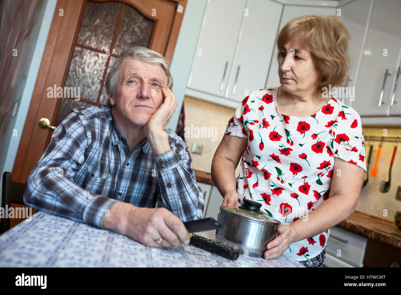 Lazy Caucasian man listens indifferently his wife, sitting with a TV remote in the kitchen Stock Photo