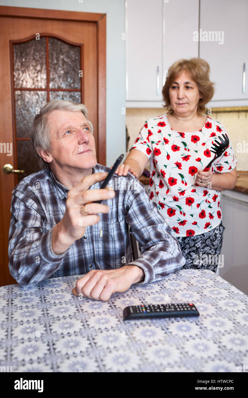 Senior wife asking lazy husband to escape from watching TV in the kitchen, mature couple relationship Stock Photo