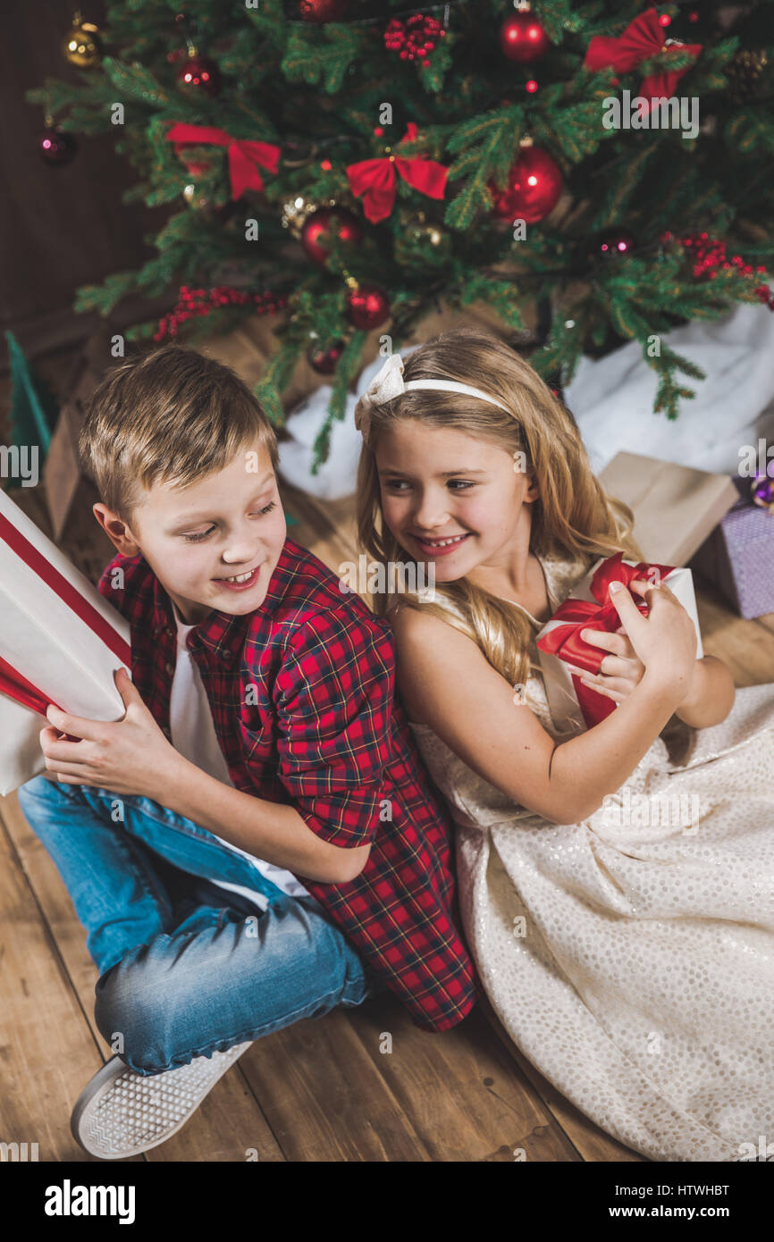 Cute happy siblings sitting near Christmas tree and holding gift boxes ...