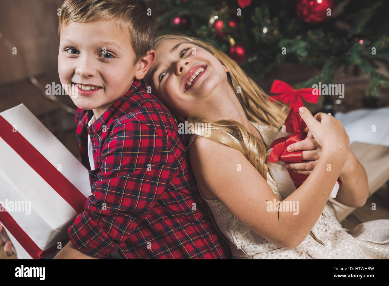 Cute happy siblings sitting near christmas tree and holding gift boxes ...