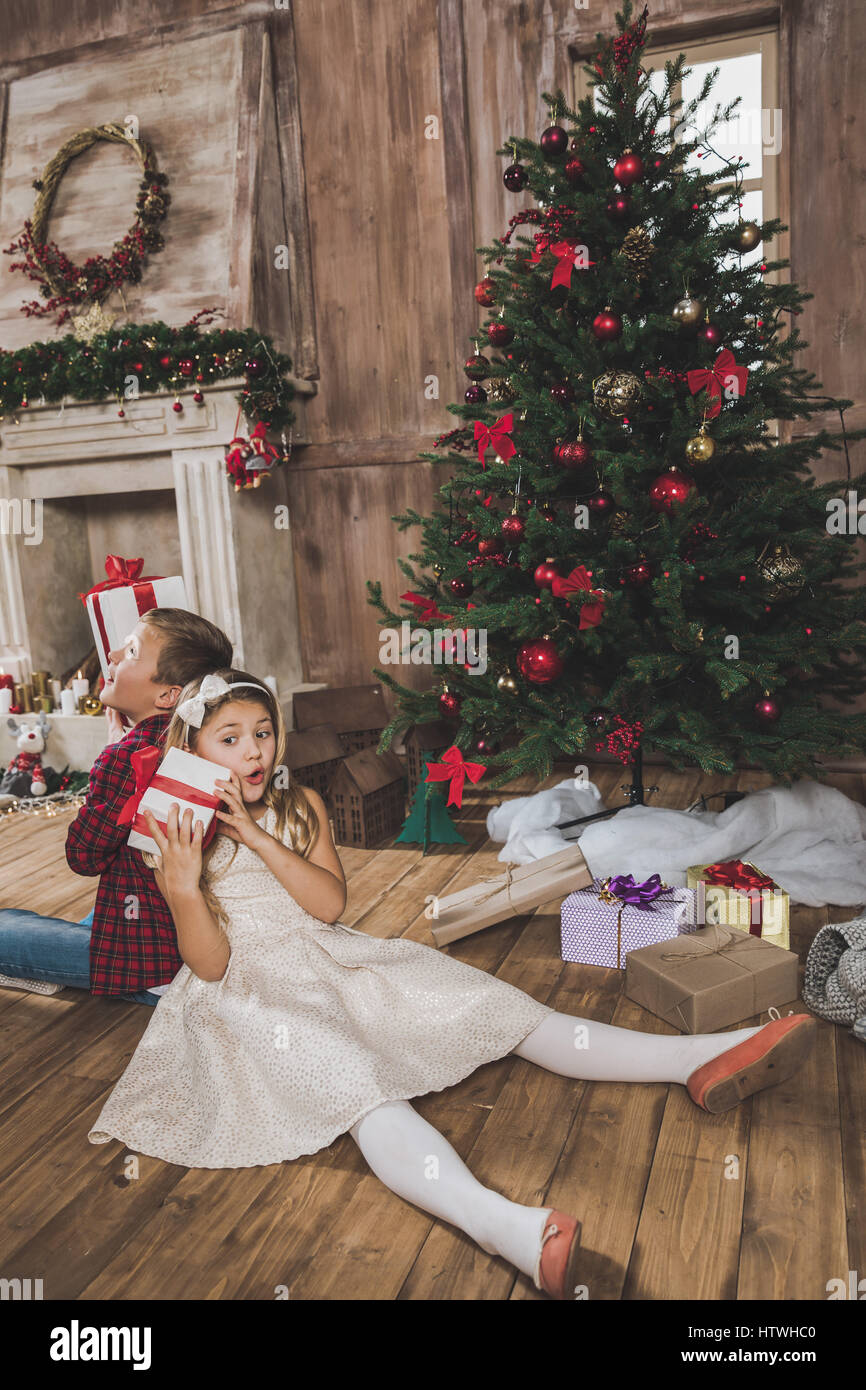 Cute happy siblings sitting near christmas tree and holding gift boxes ...