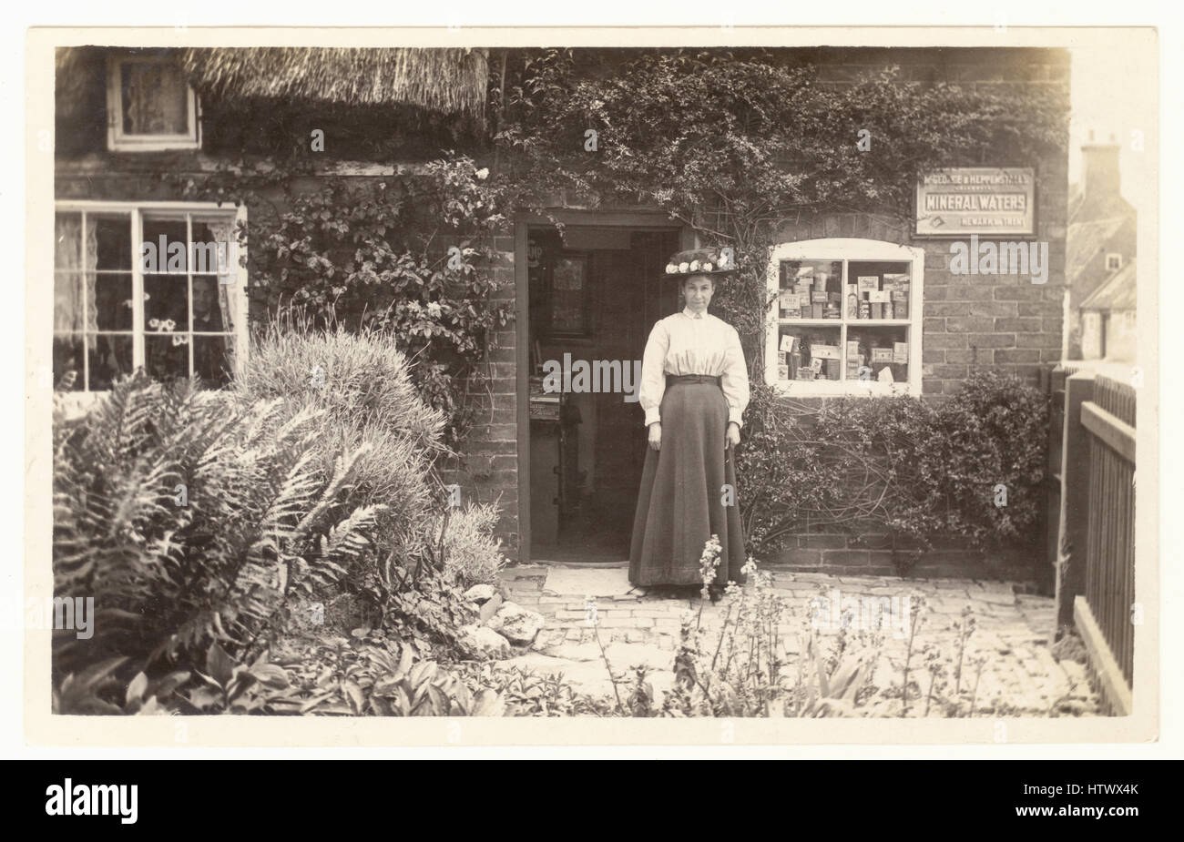 Original Edwardian nostalgic postcard of lady shopkeeper owner, sole trader, outside her rural village shop, goods in window, at her home, possibly Newark on Trent, Nottinghamshire U.K. circa 1905 Stock Photo