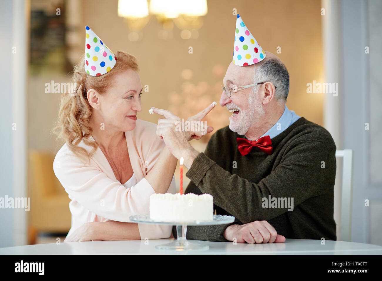 Portrait of playful senior couple sitting at table with cake and wearing party hats trying to smudge each other with cream Stock Photo