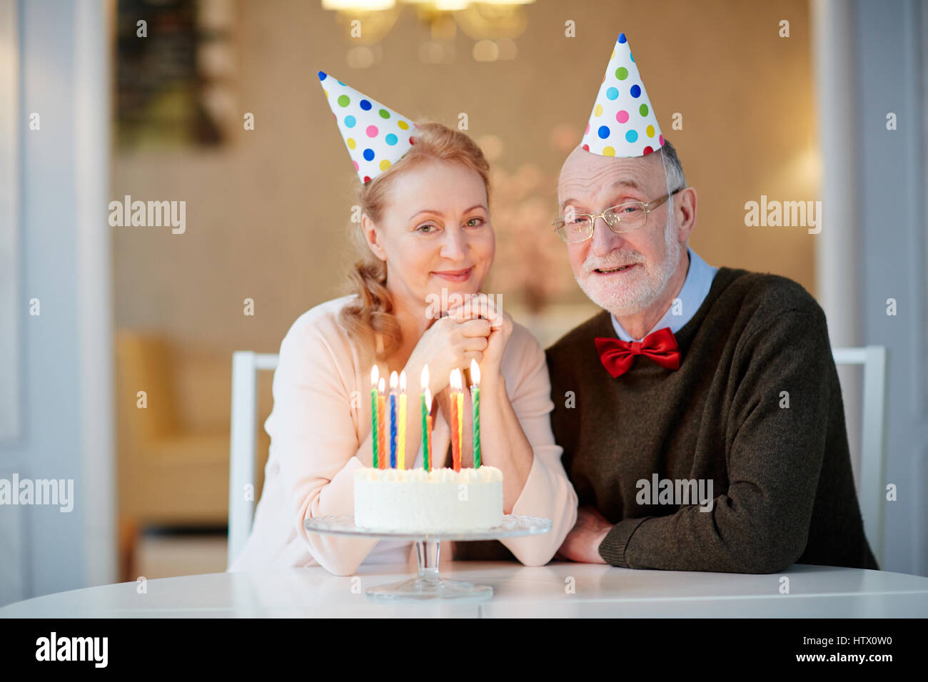Portrait of loving senior couple celebrating birthday together sitting at table with cake and wearing party hats smiling and  looking at camera Stock Photo