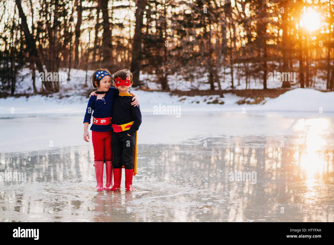 Boy and girl standing on frozen lake wearing superhero costumes Stock Photo
