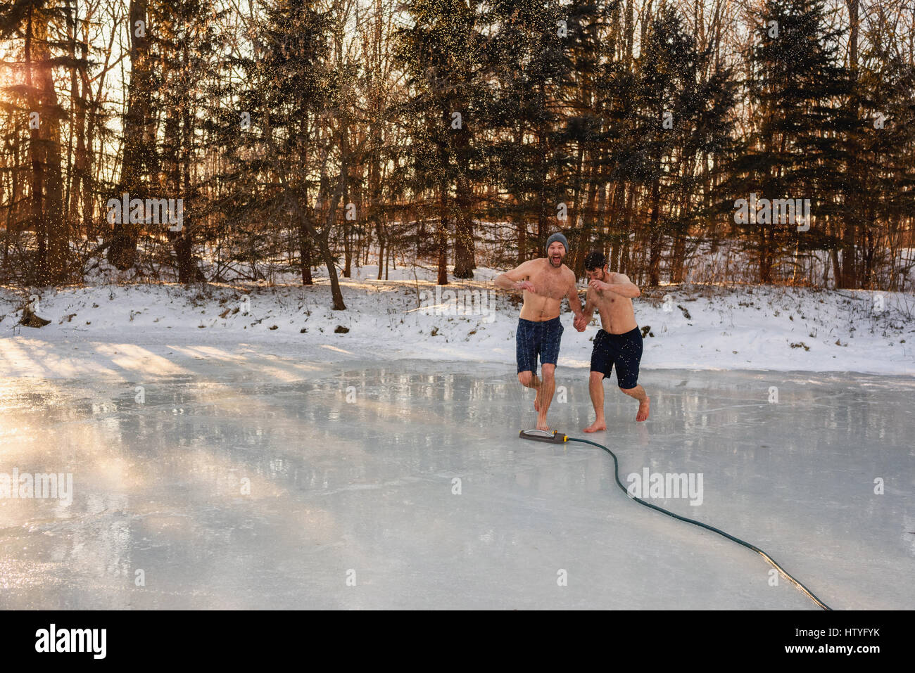 Two men in shorts running through sprinkler on frozen lake holding hands Stock Photo