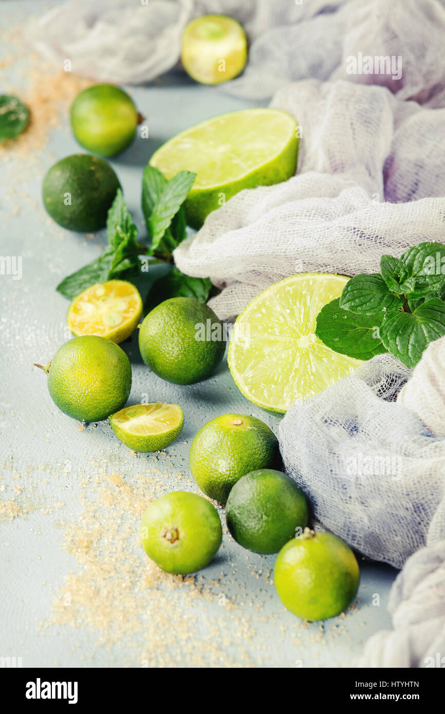 Ingredients for mojito cocktail, whole, sliced lime and mini limes, mint leaves, brown crystal sugar over gray stone texture background with gauze tex Stock Photo
