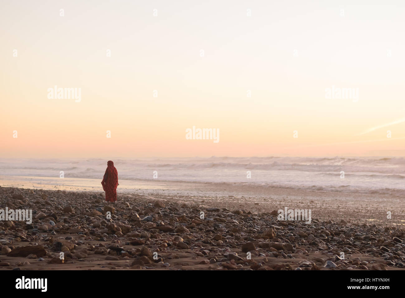 A woman in a red dress walking at the beach at sunset in Morocco. Stock Photo