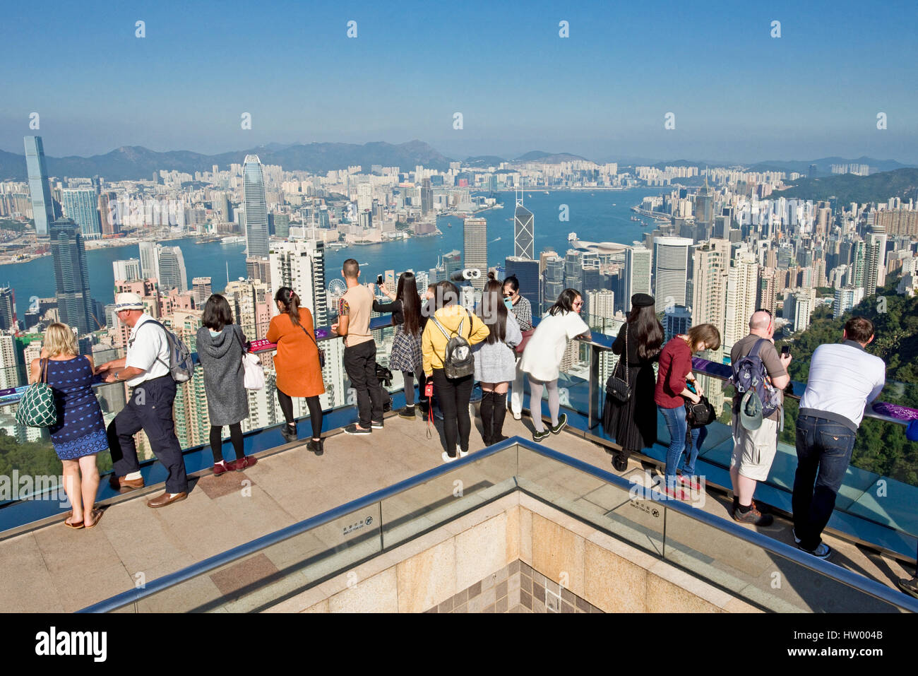 Tourists taking photographs from Sky Terrace 428 on top of the Peak Tower over Hong Kong and Victoria Bay on a clear sunny day with blue sky. Stock Photo