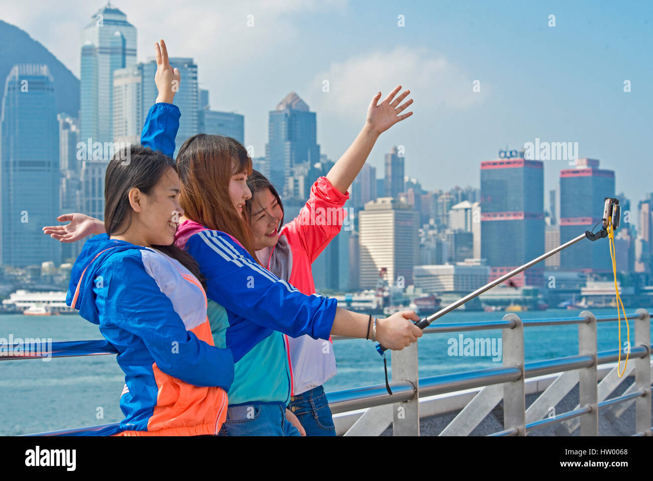 3 teenage asian girls taking a selfie stick portrait with their hands in the air smiling and laughing from the Kowloon Public Pier in Hong Kong. Stock Photo