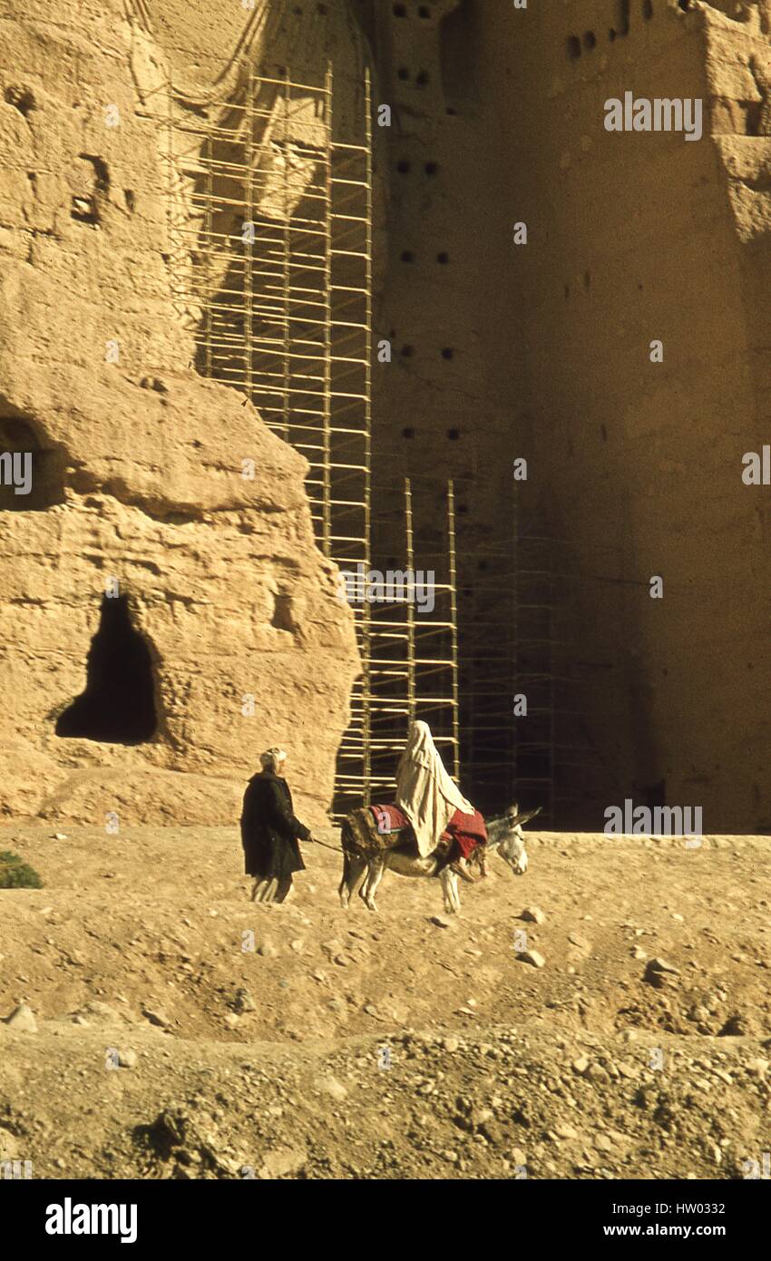 View of scaffolding attached to the base of a giant Buddha statue in Bamiyan, Hazarajat, Afghanistan, November, 1975. An Afghani man leads a donkey carrying a veiled woman in a burka past the Buddha's enormous niche. Stock Photo