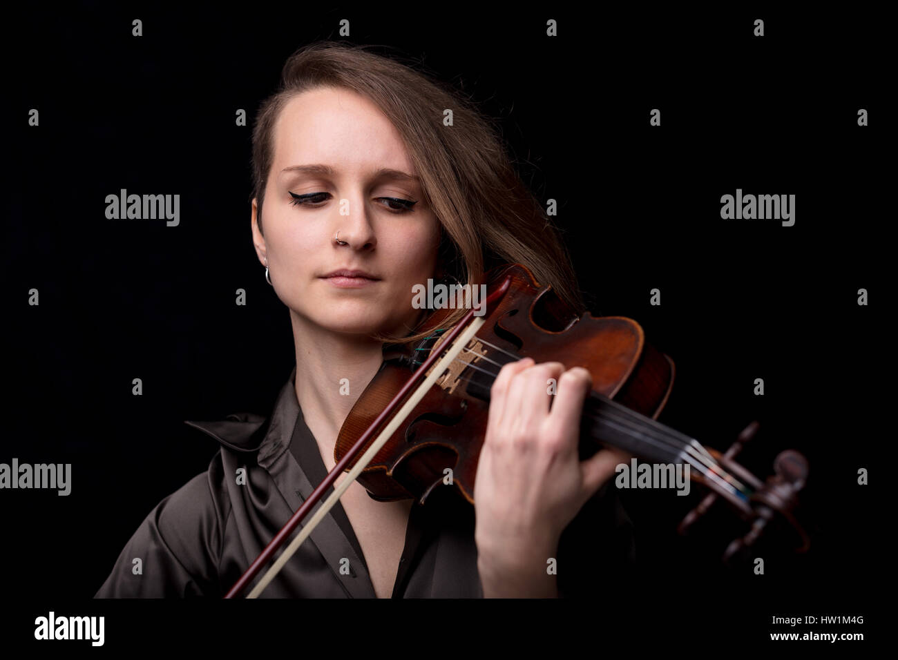 portrait of a young beautiful woman violinist player playing her instrument on her shoulder holding bow. portrait in a blurred dark room in background Stock Photo