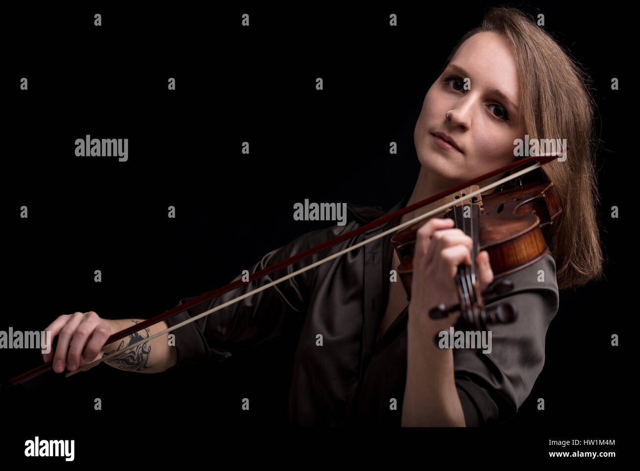 Young beautiful woman violinist player  looking at camera over instrument on her shoulder holding bow. Portrait in a blurred dark room in background.  Stock Photo