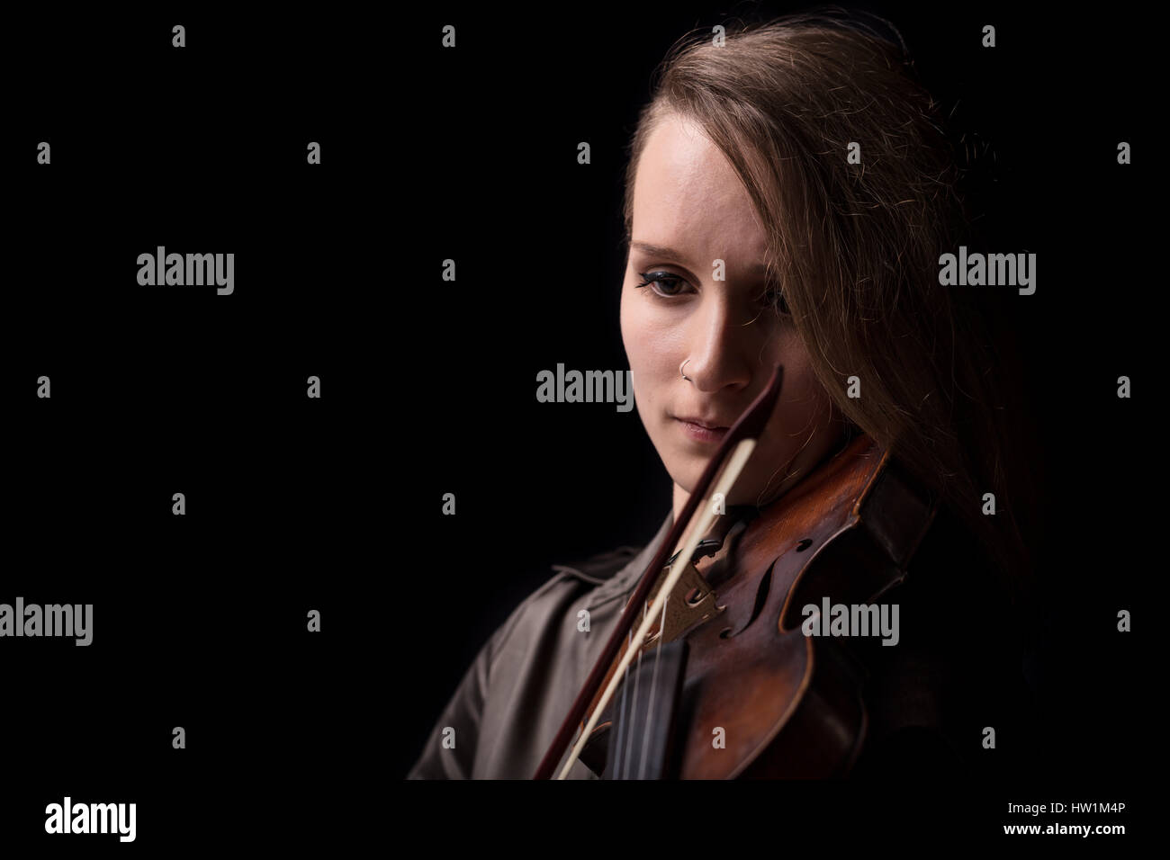 Young beautiful woman violinist player playing her instrument on her shoulder holding bow. portrait in a blurred dark room in background. Concept of c Stock Photo