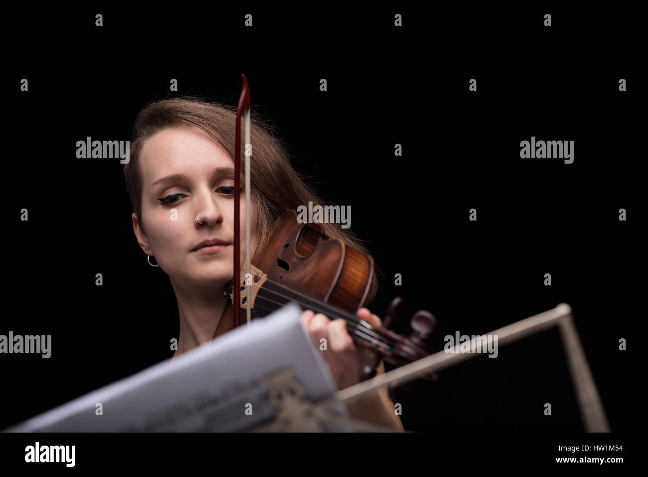 Young beautiful woman violinist player playing her instrument on her shoulder holding bow. portrait in a blurred dark room in background. Concept of c Stock Photo