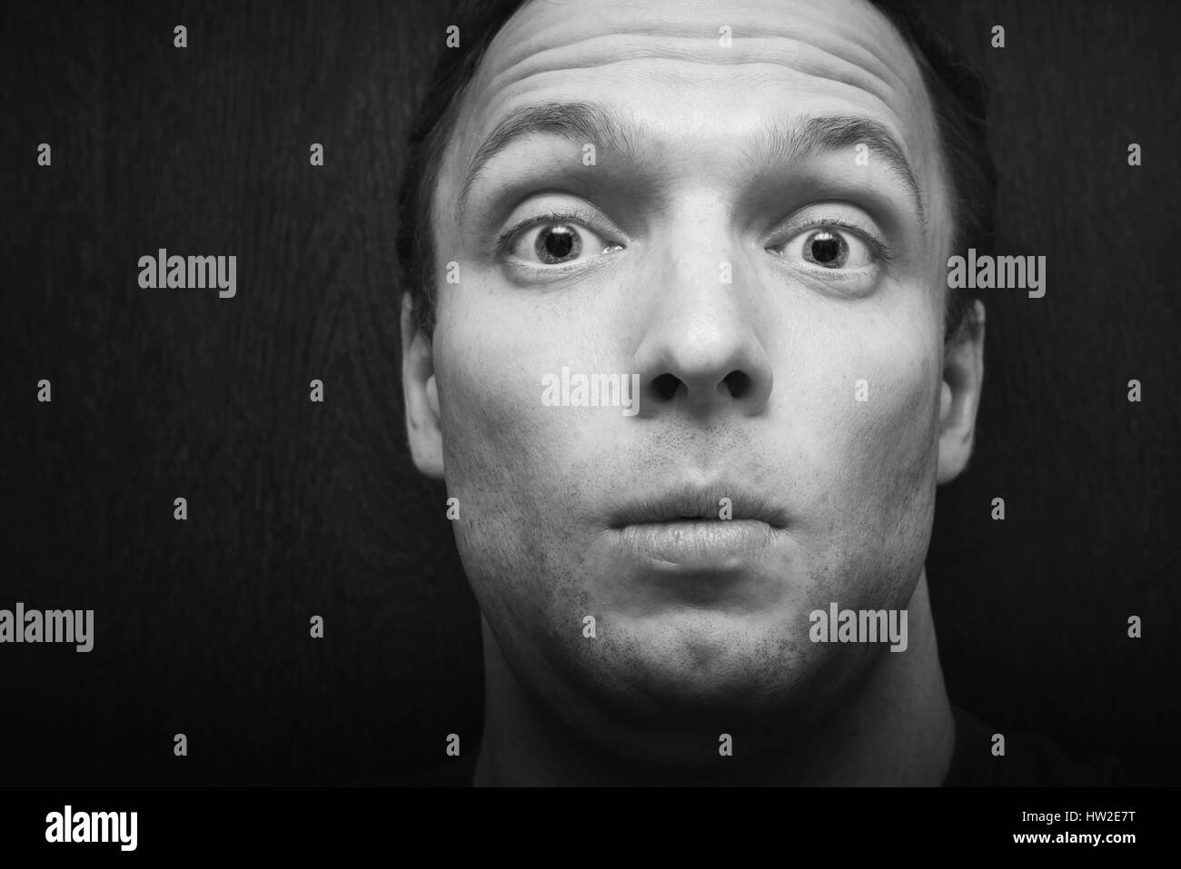 Young surprised Caucasian man. Close-up studio face portrait over dark wooden wall background, selective focus, black and white photo Stock Photo