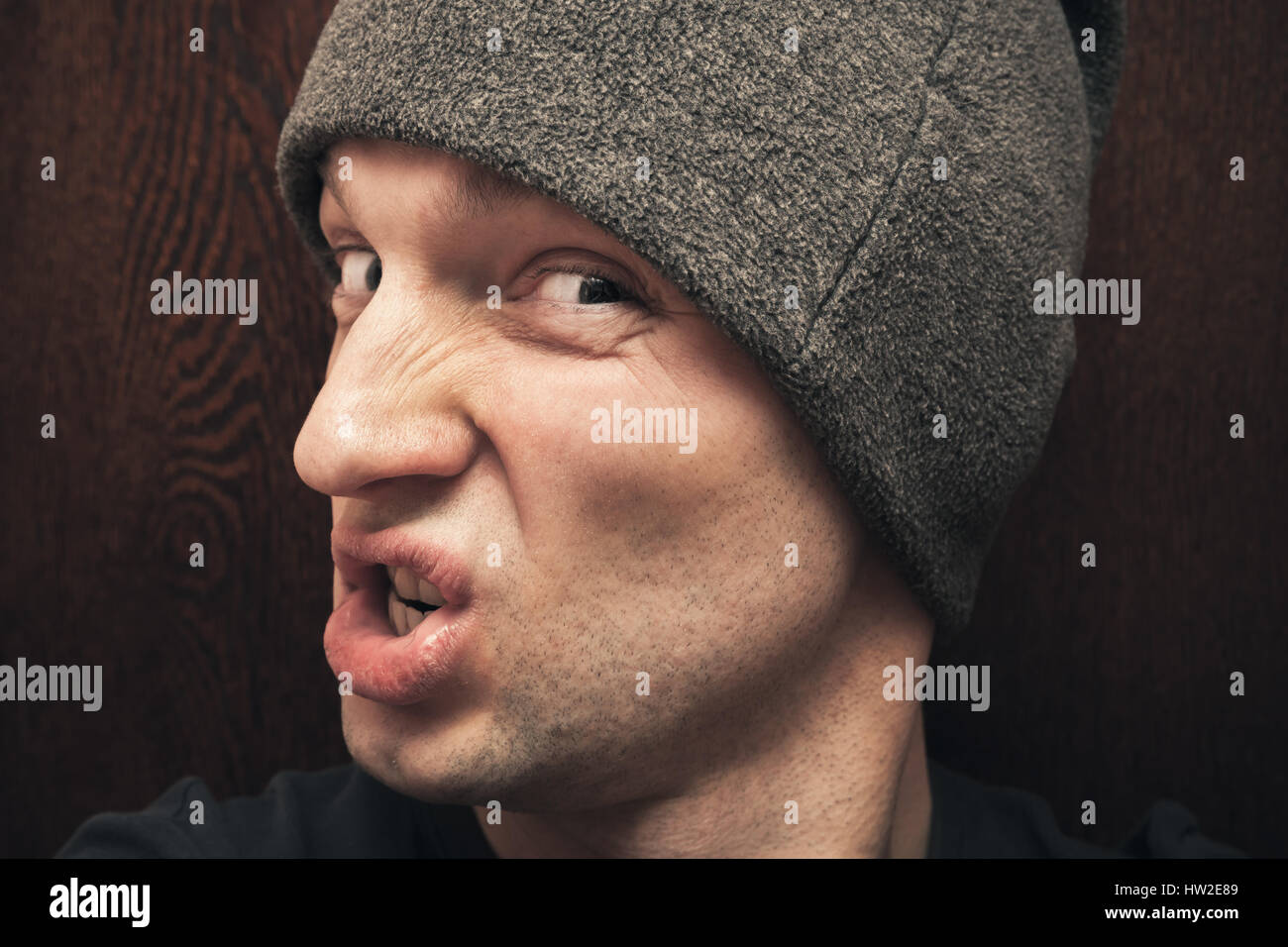 Young agressive Caucasian man in gray hat. Close up studio face portrait on dark wooden wall background, selective focus Stock Photo