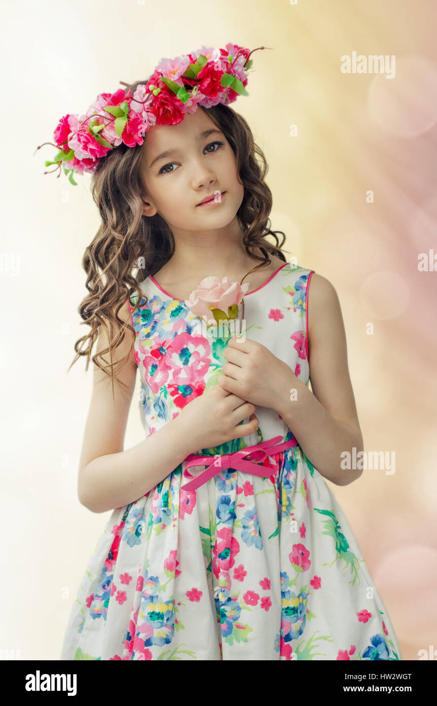 Portrait of cute little girl in nice spring dress, with flower wreath on head, holds pink rose in hands Stock Photo