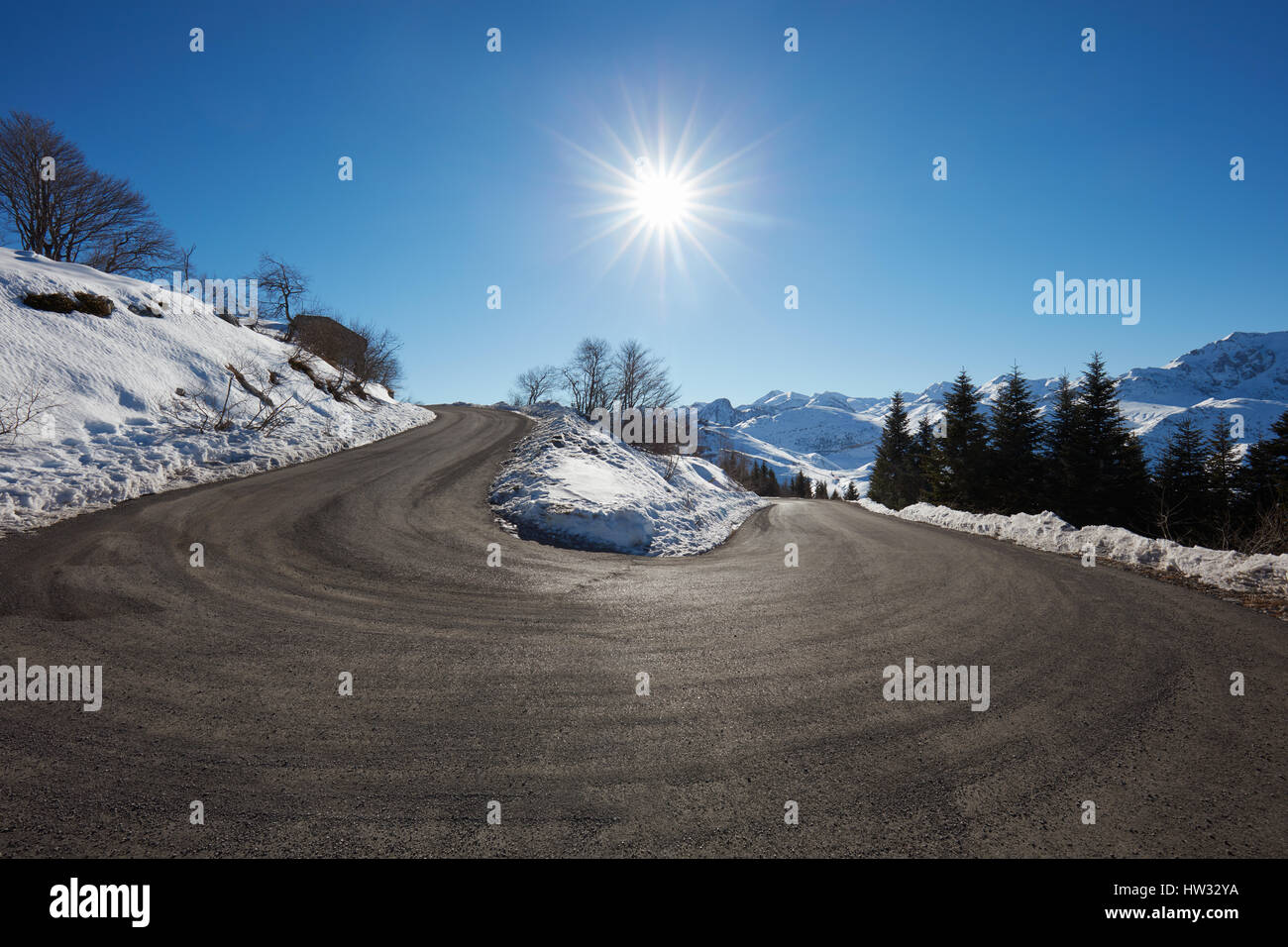 Large empty mountain road curve on Alps with snow on sides, blue sky and sun Stock Photo