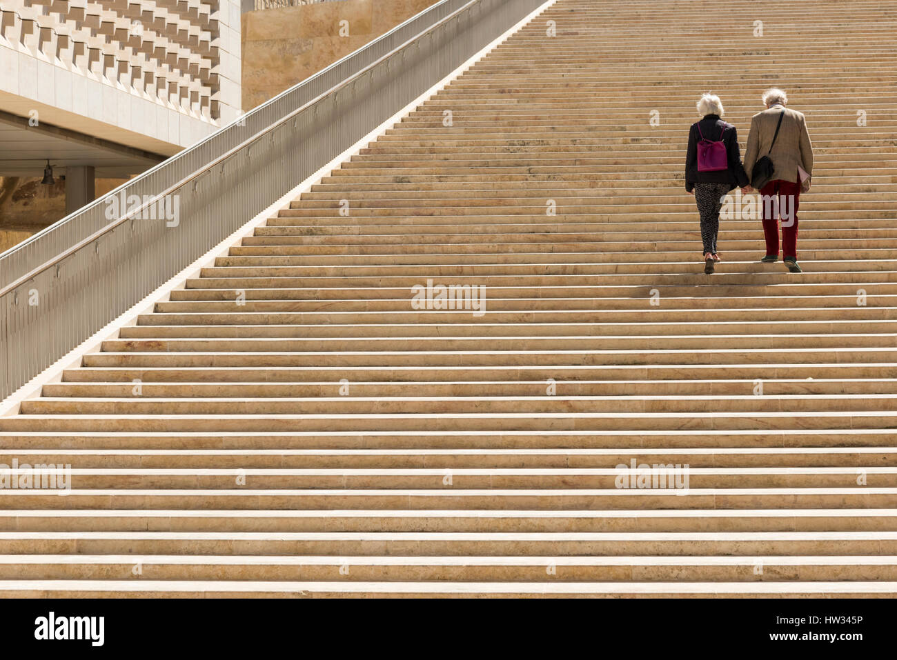 Two older people walking hand in hand together up the stone steps of the Renzo Piano designed entrance and parliament building at the City of Valetta  Stock Photo