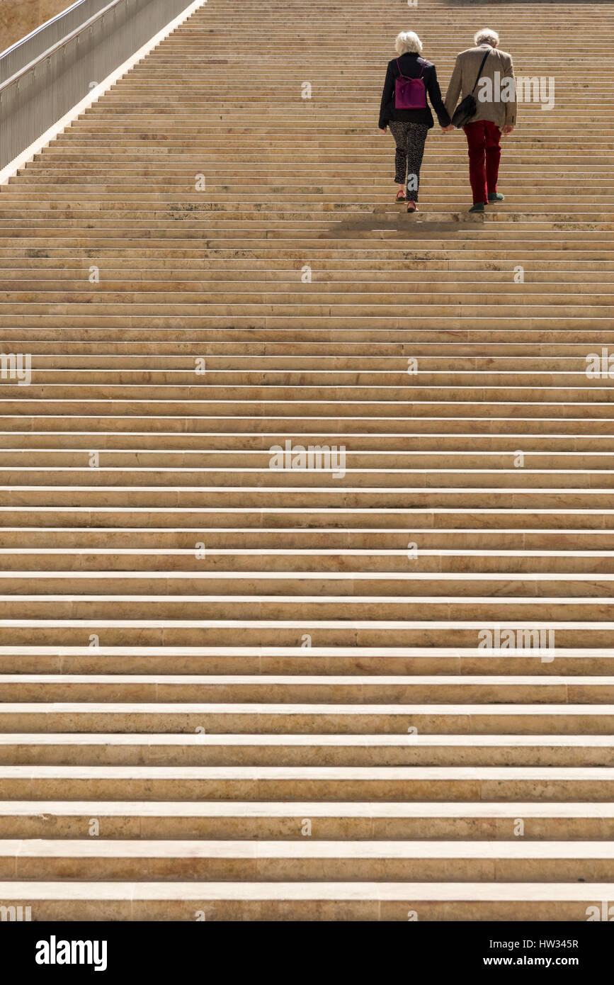 Two older people walking hand in hand together up the stone steps of the Renzo Piano designed entrance and parliament building at the City of Valetta  Stock Photo