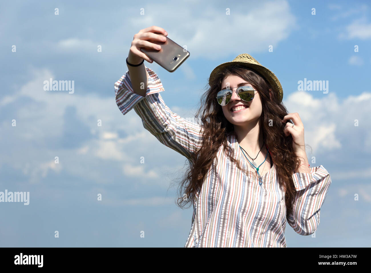 Girl hippie Style Clothing casual Shirt Hat Sunglasses and other accessories taking self portrait photo on mobile telephone camera on blue sky and sum Stock Photo