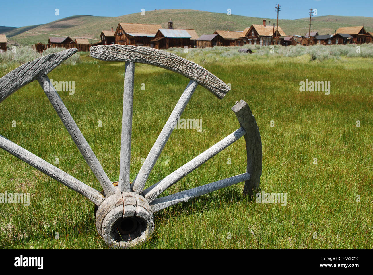 A wooden wagon wheel stuck in the ground in the foreground and beyond is a row of several old buildings of Bodie, a ghost town in California. Stock Photo