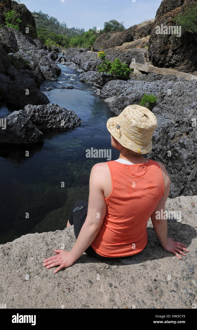 A mid-adult woman with her back to the camera sits on a rock above a river looking off to the side. She is wearing a peach tank top and a straw hat. Stock Photo