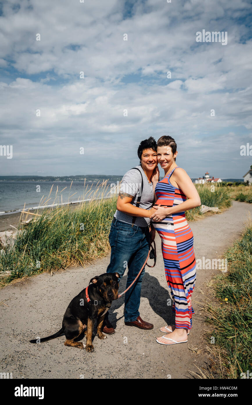 Portrait of pregnant lesbian couple with dog hugging at waterfront Stock Photo