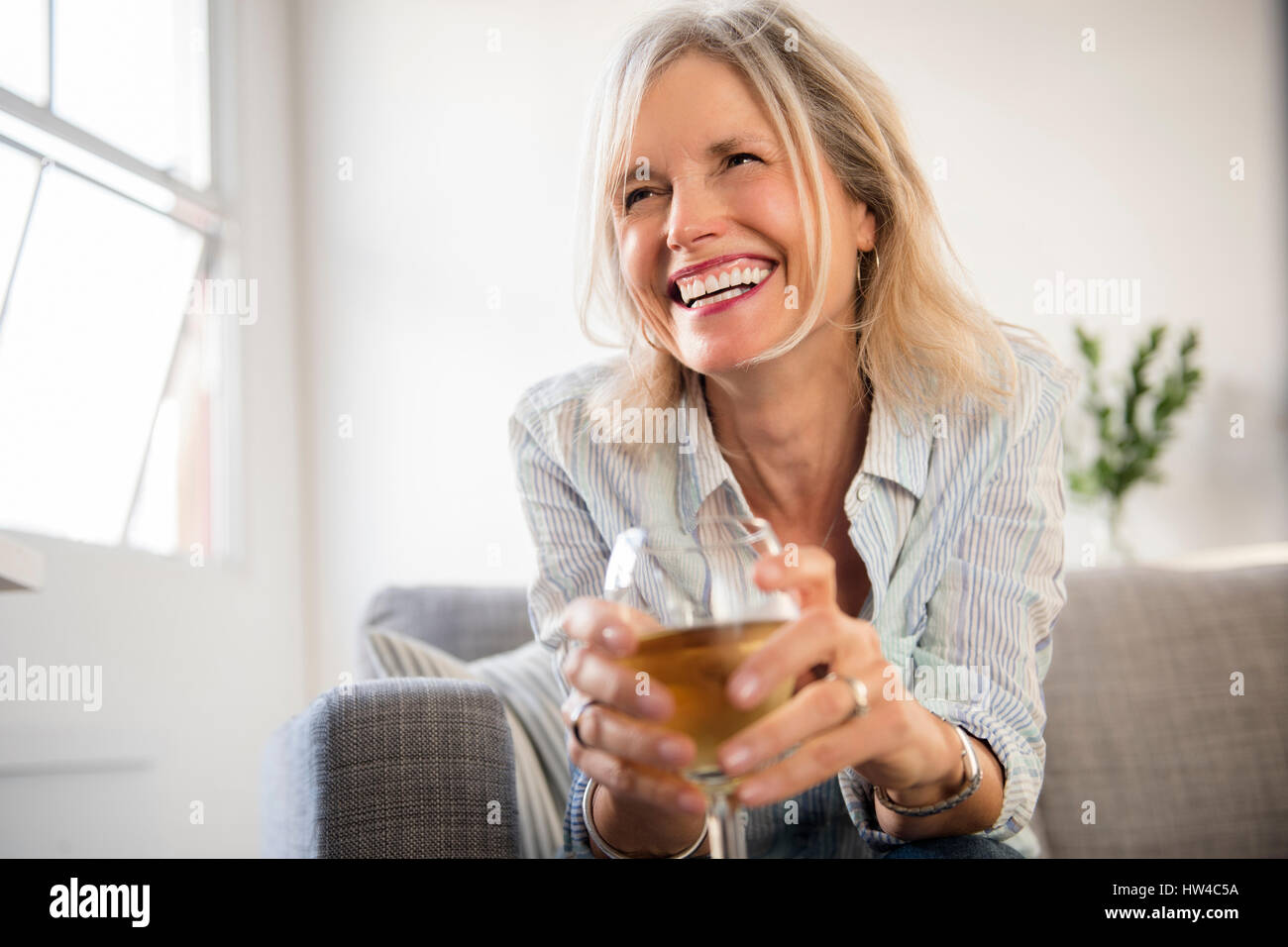 Smiling Caucasian woman sitting on sofa drinking white wine Stock Photo