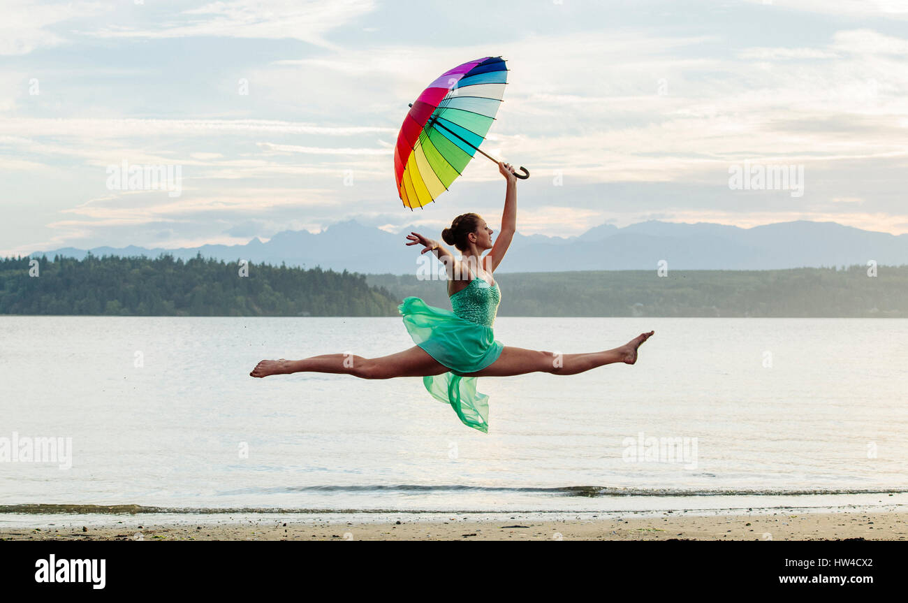 Caucasian ballerina jumping with multicolor umbrella on beach Stock Photo