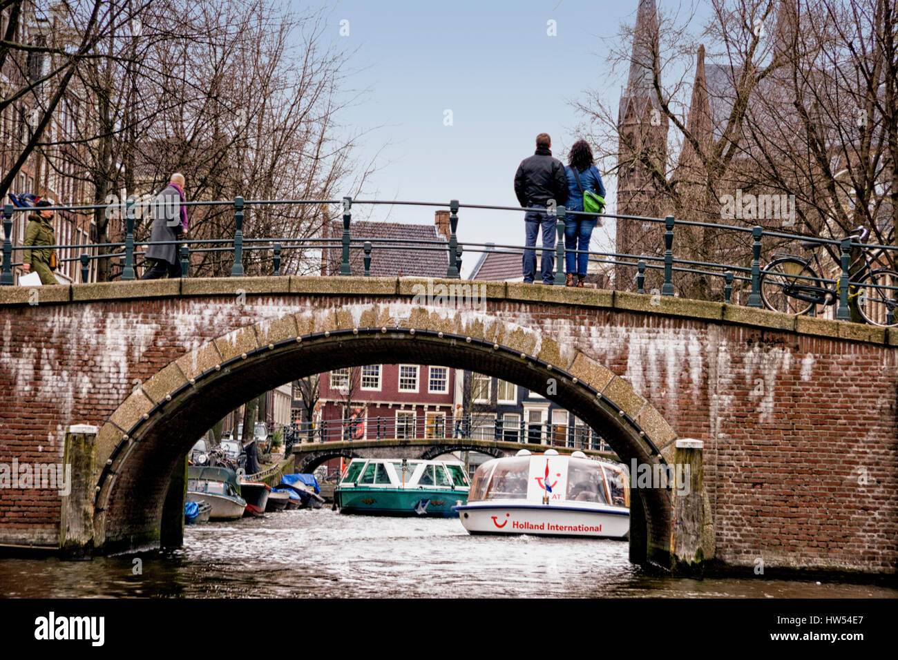 Tourists standing on a bridge, watching canal boats. Amsterdam, Netherlands. Stock Photo