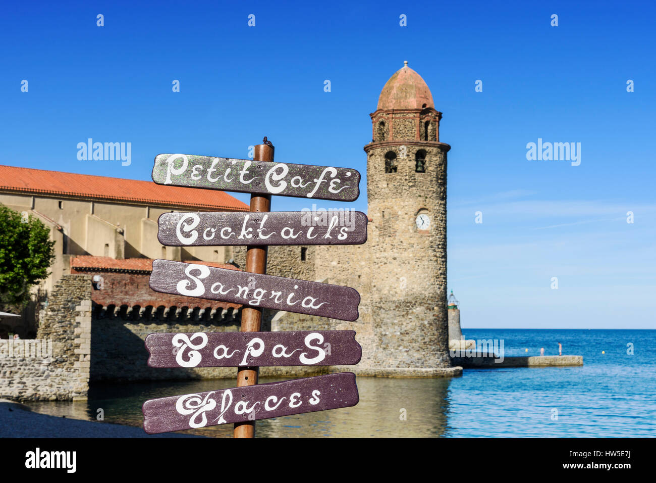 Focus on a cafe sign at a beachside cafe overlooked by the bell tower of the Church of Notre Dame des Anges, Collioure, Côte Vermeille, France Stock Photo