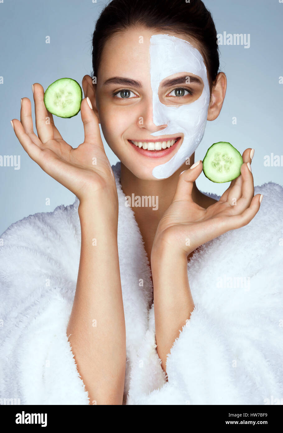 Laughing beautiful model with the slices of cucumber in hands. Photo of girl in white bathrobe and with moisturizing facial mask. Grooming himself Stock Photo