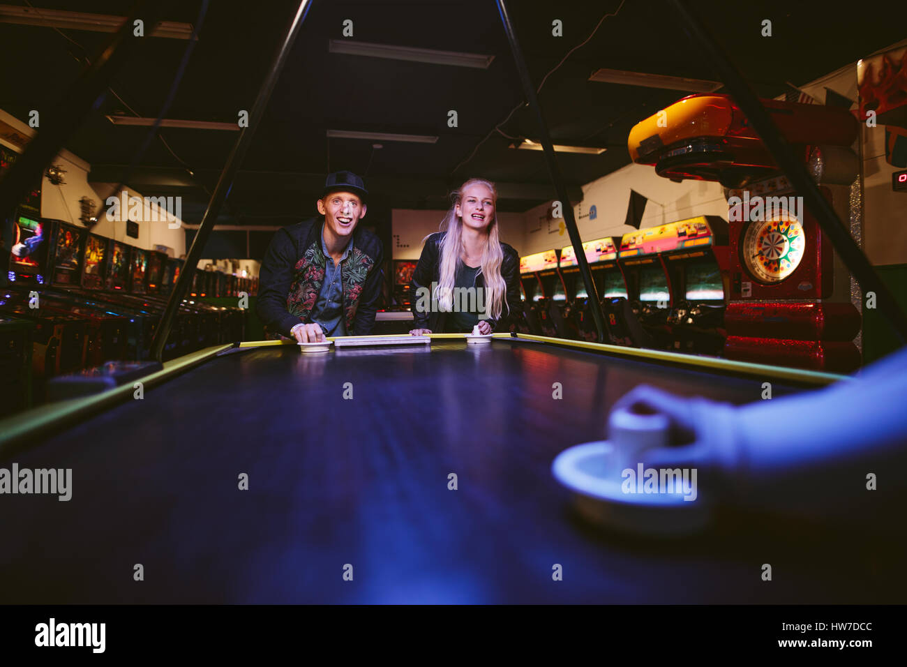 Smiling young friends playing air hockey game. Man and woman playing a game of air hockey in the game room at amusement park. Stock Photo