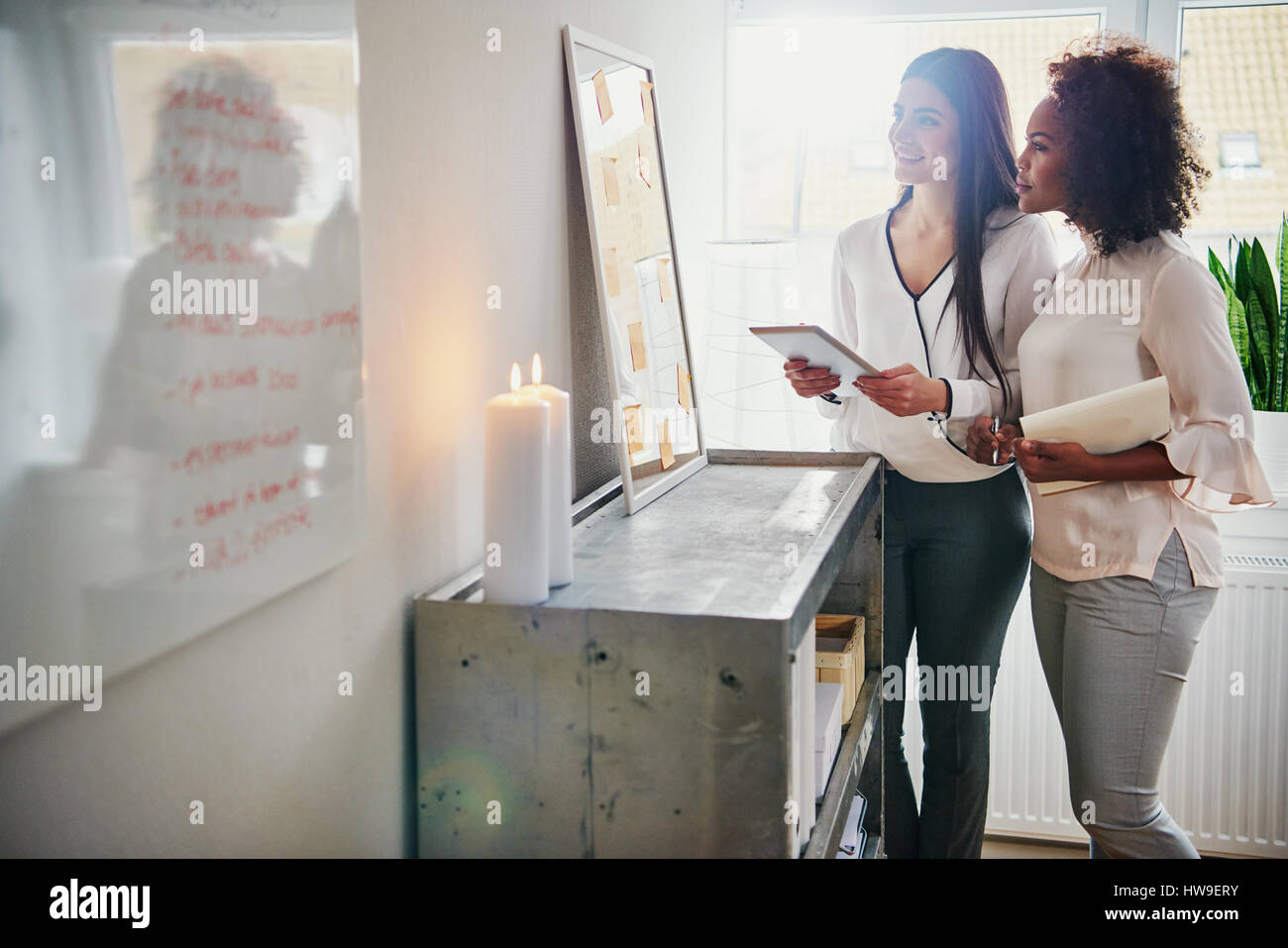 Two businesswomen holding a tablet and paperwork standing looking at their memo board with new ideas and planning in the office as they discuss their Stock Photo