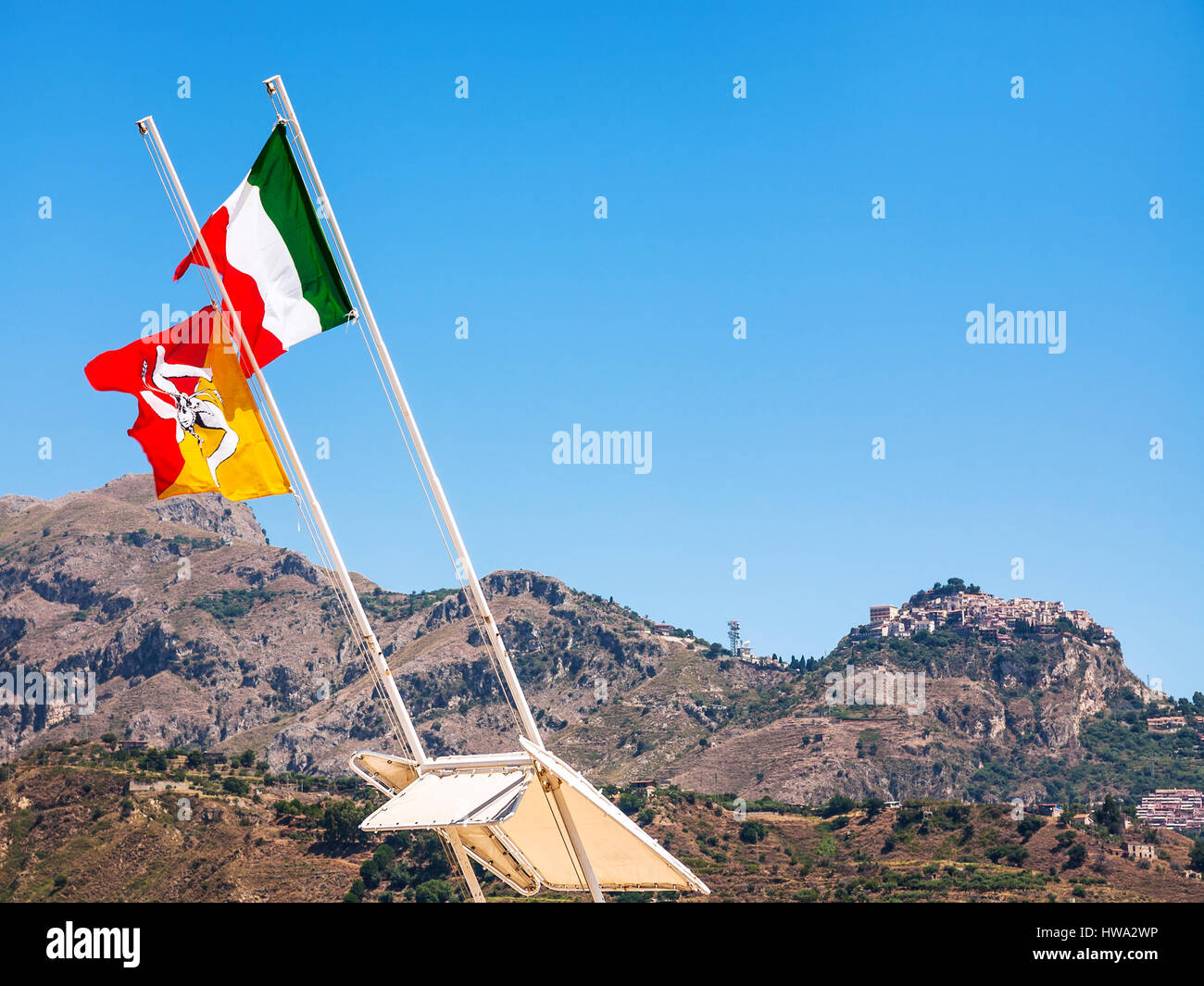 travel to Italy - Italian and Sicilian flags flutter by wind and view of Taormina town on mountain in Sicily Stock Photo