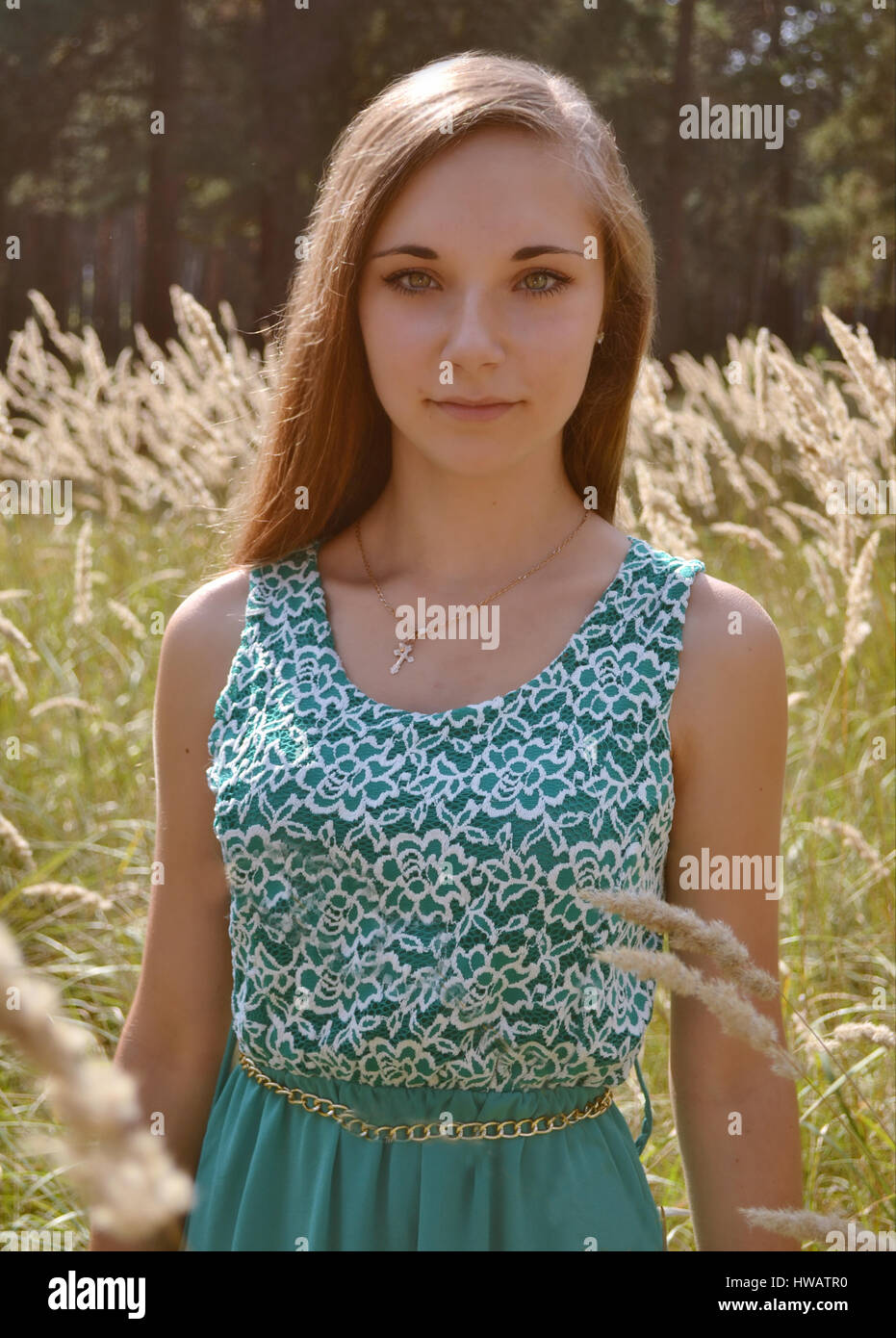 Beautiful girl with long hair, wearing blue, green dress outdoors standing in the middle of the sunny clearings full of flowers, ears, wheat in forest Stock Photo