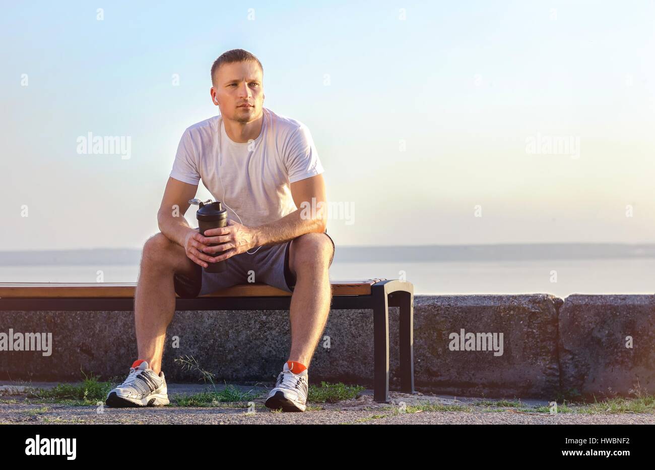 young athlete of Caucasian appearance with a shaker for a protein cocktail or water in his hands, sits on a bench after training in the open air. Stock Photo