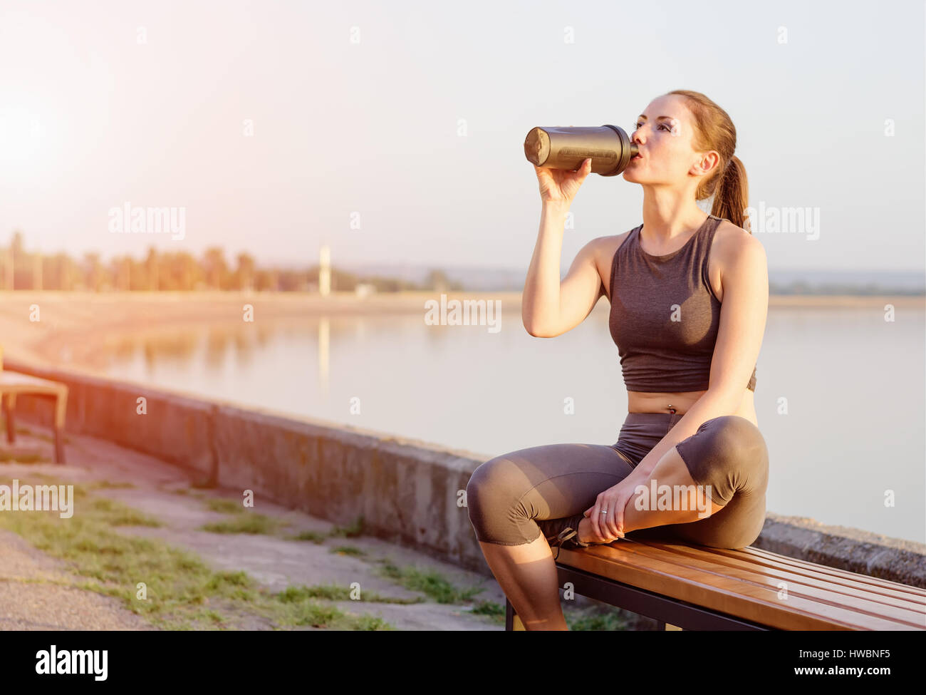 young girl of Caucasian appearance drinks a protein cocktail from a shaker after jogging in the open air. The bright sun illuminates it. Stock Photo
