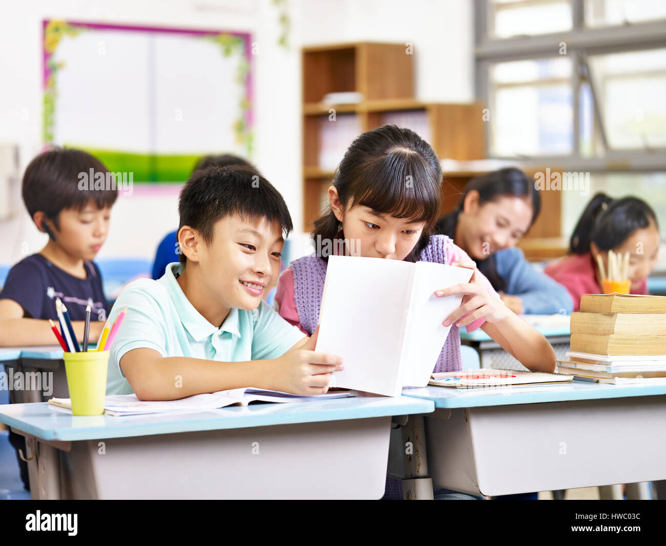 asian elementary schoolgirl and schoolboy sharing a book in classroom. Stock Photo