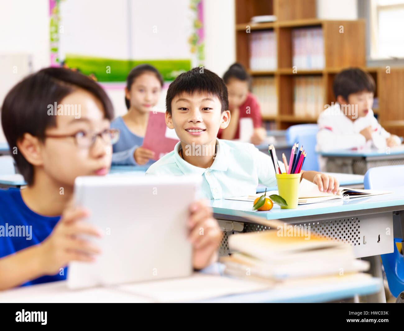 asian elementary schoolboy looking with curiosity at a tablet computer held by his classmate, focus on the boy in background. Stock Photo
