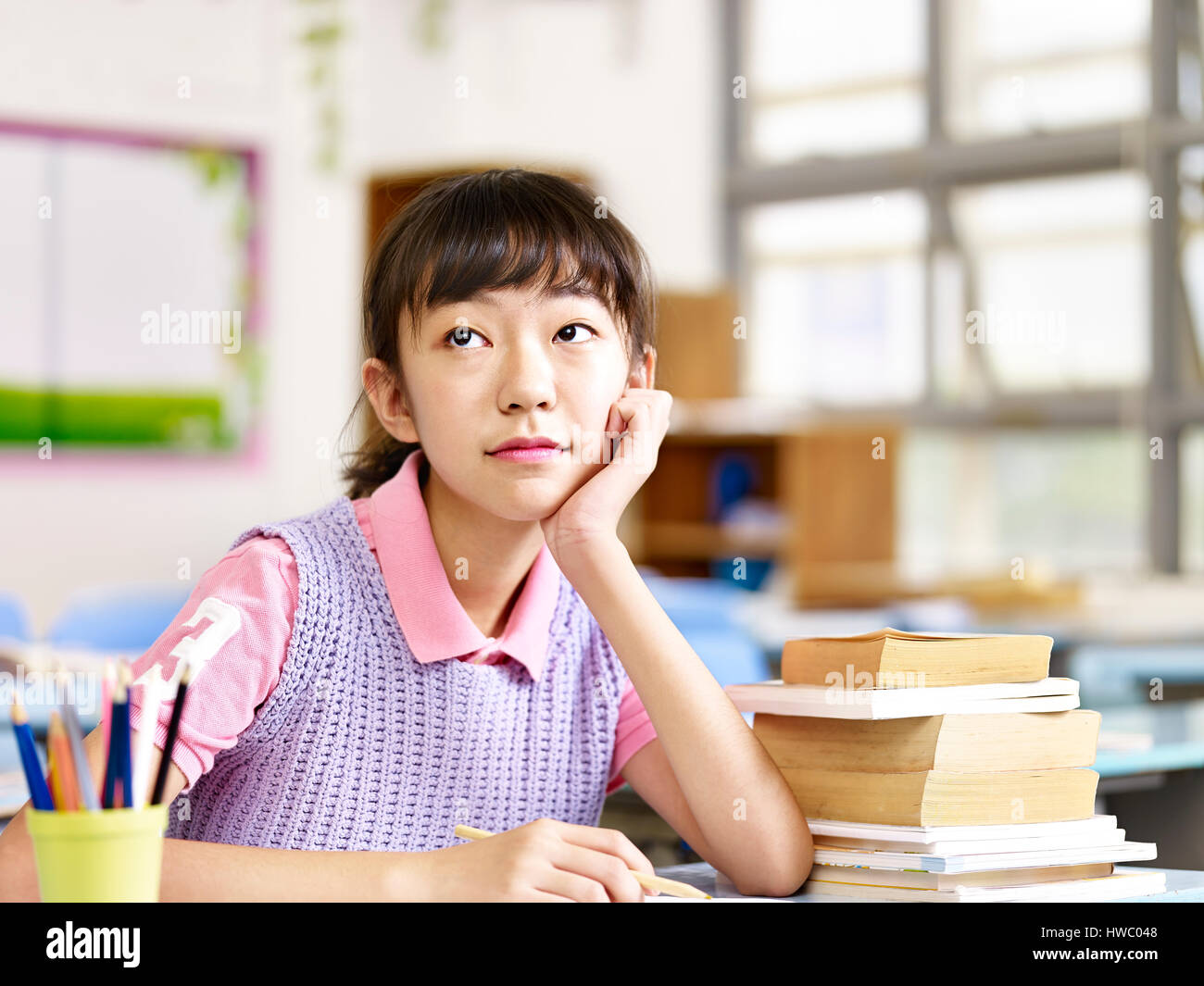 asian elementary schoolgirl sitting at desk looking up thinking in classroom. Stock Photo