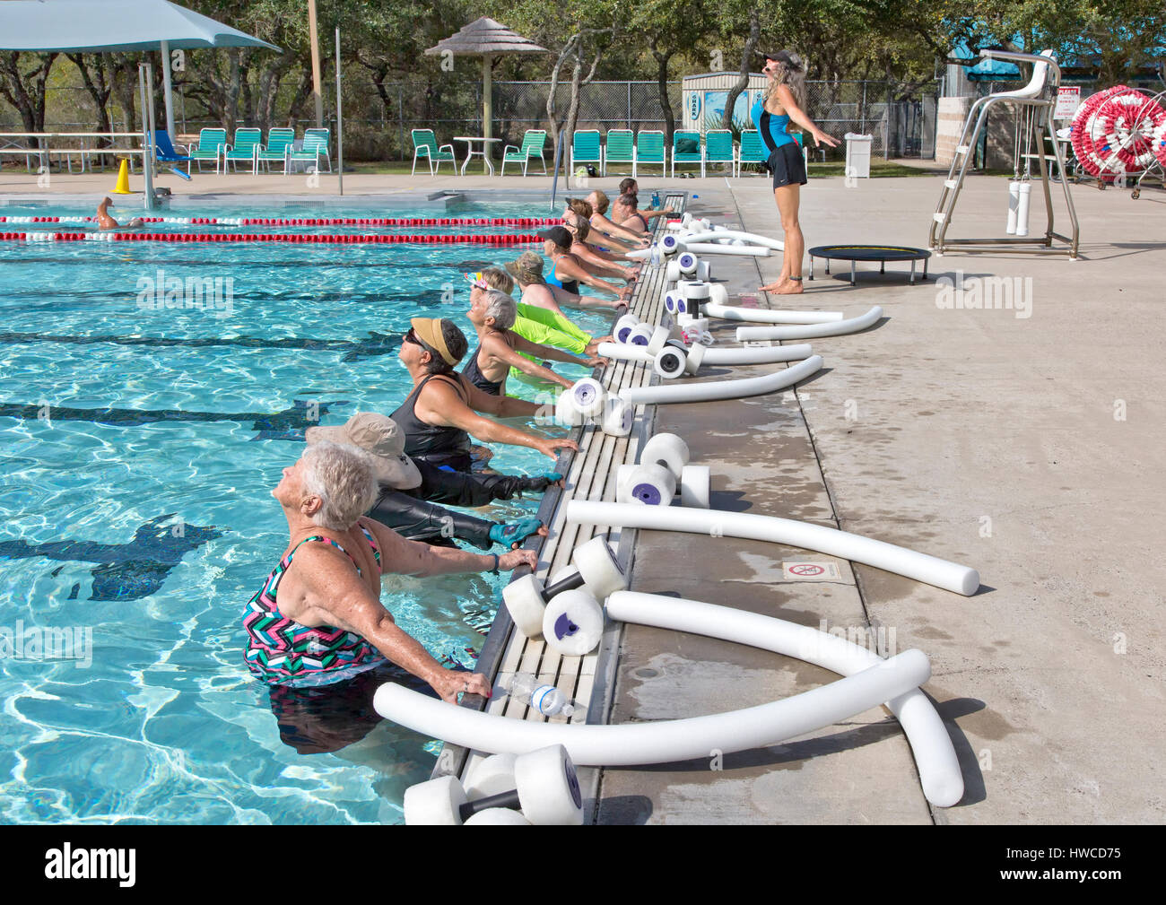 Group of women (various ages) attending Water Aerobics Class,  instructor demonstrating 'proper stretch exercise', styrofoam dumbbells & water noodles. Stock Photo