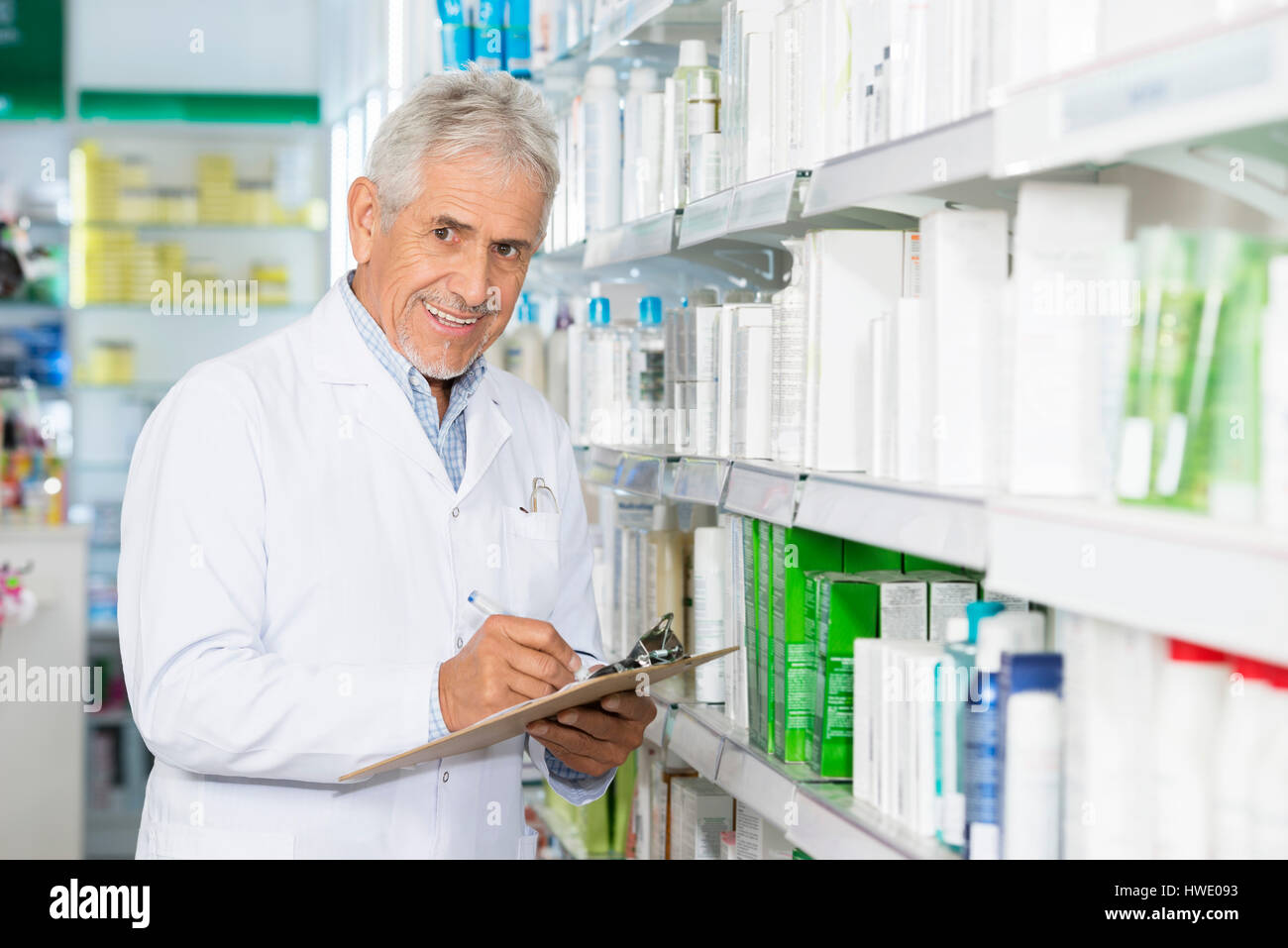 Smiling Chemist Writing On Clipboard While Standing By Shelves Stock Photo