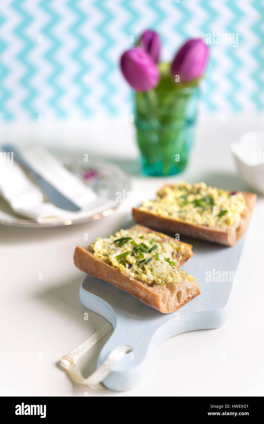 Close-up of fresh baguette sandwich spread with with egg salad on blue cutting board with bunch of tulips in background Stock Photo
