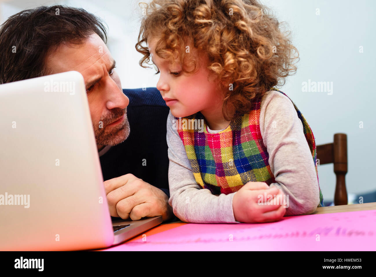 Girl peering at father's laptop at table Stock Photo